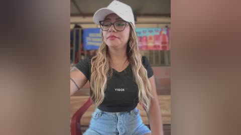 Video of a young woman with wavy, blonde hair, wearing a black t-shirt with \"VIGO\" and blue jeans, sitting in a brick-walled room with colorful posters in the background.