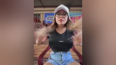 Video of a young woman with glasses, light skin, and brown hair, wearing a black crop top and high-waisted jeans, seated in a booth, blurred background.