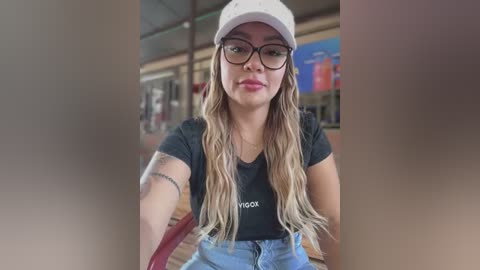 Media: Video of a young Latina woman with long, wavy blonde hair, wearing a white baseball cap, black glasses, and a black t-shirt with \"H&M\" text. She has a tattoo on her left arm and is seated in a modern, well-lit indoor setting.