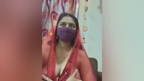 Media: Video of a South Asian woman with medium skin tone, wearing a red saree, purple face mask, and a gold necklace. Background includes a curtain with a floral pattern, dreamcatcher, and a white wall.