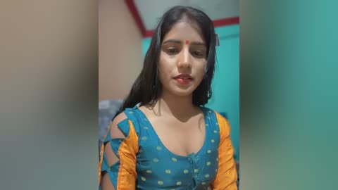 Media: Video of a young South Asian woman with medium skin tone, wearing a blue top with yellow polka dots and orange sleeves, with a red bindi on her forehead, in a teal-colored room.