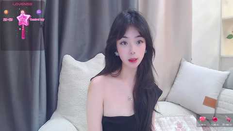 Media: Video of an East Asian woman with long black hair, fair skin, and red lipstick, sitting on a beige sofa in a modern living room with grey curtains.