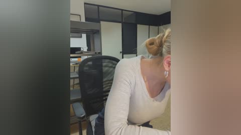 Media: A video shows a woman with blonde hair in a bun, wearing a white top, seated at a desk in an office with black-framed windows and beige walls.