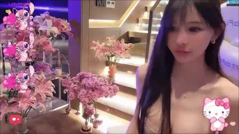 Media: Video of an Asian woman with long black hair and fair skin, wearing a sheer top, standing next to a shelf of pink flowers. The background features a modern staircase and a My Melody figurine.