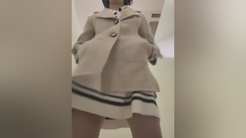 Video of a woman in a cream coat and white dress with navy stripes, standing in a narrow hallway with beige walls. Her hands are in her coat pockets.