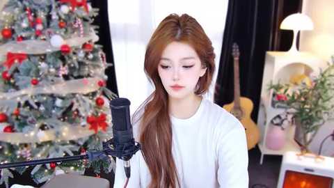 Media: Video of a young Asian woman with long, straight brown hair, singing into a microphone while standing in a brightly lit, festive room with a decorated Christmas tree and a guitar in the background.