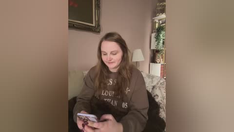 Media: Video of a young Caucasian woman with long brown hair, wearing a dark brown sweater, sitting on a bed, reading a magazine in a cozy, dimly lit room.