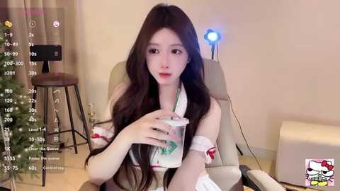 Media: Video of an East Asian woman with long, wavy black hair, fair skin, and red lipstick, seated in a beige chair, holding a green and white drink, in a room with a Christmas tree, and a small blue light.