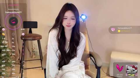 Media: Video of a young Asian woman with long, straight black hair, wearing a white robe, sitting on a chair in a cozy room with a lit lamp, displaying a social media livestream interface.