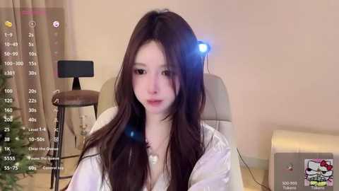 Video of a young East Asian woman with long, dark brown hair, pale skin, and soft facial features, wearing a white robe. She sits indoors, with a digital camera on a tripod, and a small table and chair visible.