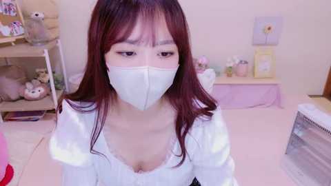 Media: Video of an Asian woman with long, straight, brown hair, wearing a white face mask, white dress, and sitting in a pastel-colored room with soft lighting.