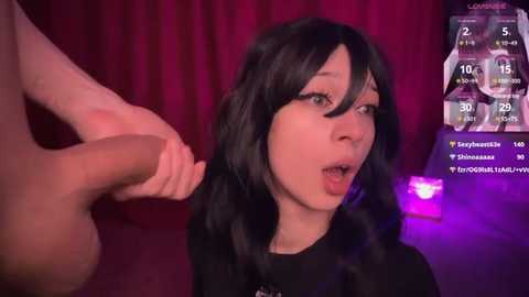 A video of a young Asian woman with long black hair, wearing a black top, engaged in a live stream. She is looking at a large, erect penis held by a man, her mouth open in surprise.