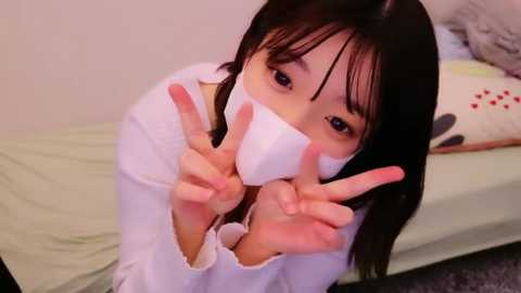 Media: Video of an Asian woman with straight black hair, wearing a white face mask, and making peace signs with both hands, kneeling on a green bed with a pillow.