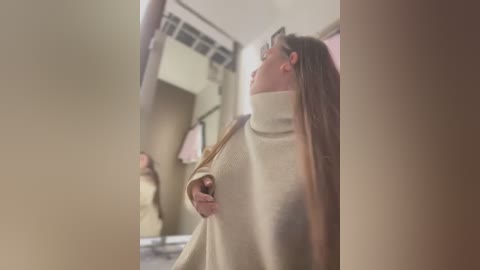 Video of a woman with long brown hair, wearing a beige turtleneck sweater, looking upward in a dimly lit room with beige walls and a mirrored surface.