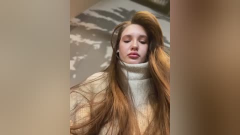 Media: Video of a young woman with long, flowing red hair, wearing a cream-colored turtleneck sweater, standing in a dimly lit room with a textured gray wall.