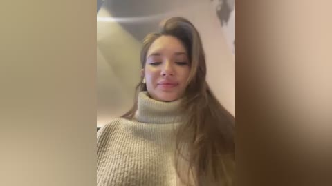 Media: Video of a young woman with long, straight brown hair styled voluminously on one side. She has fair skin, closed eyes, and wears a cream-colored turtleneck sweater. The background is blurred, suggesting a soft-focus effect.