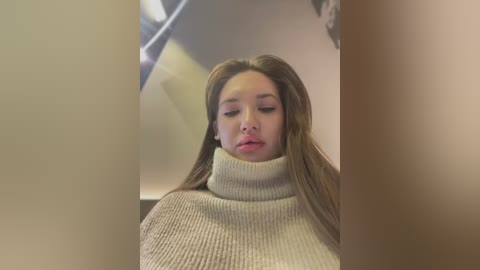 Video of a young woman with long, straight brown hair, wearing a beige turtleneck sweater, with a blurred, beige background and a faint reflection of a fan.