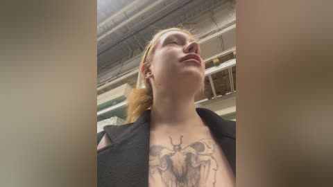 Video of a blonde woman with a large, detailed tattoo of a bee on her chest, wearing a black jacket, standing in an industrial warehouse with exposed pipes and shelves in the background.