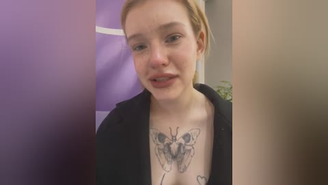 Video of a young woman with fair skin and light brown hair, wearing a black jacket, showcasing a detailed butterfly tattoo on her chest. Background includes a purple wall and blurred figures.