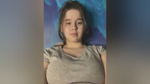 Media: Video of a young Caucasian girl with fair skin, brown hair in a messy ponytail, and a light gray T-shirt. She is lying on a blue and purple floral background, looking directly at the camera with a neutral expression.