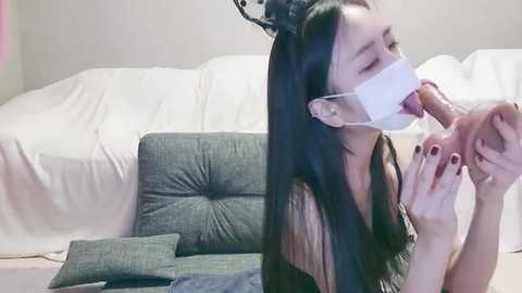 Media: A video of a young Asian woman with long black hair, wearing a white surgical mask, kneeling on a beige carpet, performing oral sex on a large dildo, set against a white couch and grey cushions.