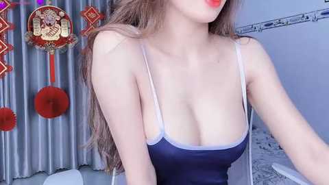 Media: Video of a fair-skinned woman with long, wavy brown hair, wearing a revealing navy blue tank top, against a backdrop of decorative red and gold Chinese New Year banners and white curtains.