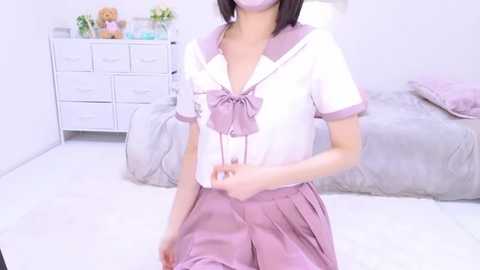 Media: Video of an Asian woman in a Japanese schoolgirl cosplay outfit, wearing a white sailor dress with a purple bow and pleated skirt. She stands in a minimalist, pastel-colored bedroom with a white dresser and teddy bear.