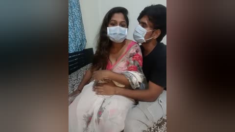 Media: Video of a woman in a pink saree and face mask sitting on a sofa with a man, also wearing a face mask, in a domestic setting.