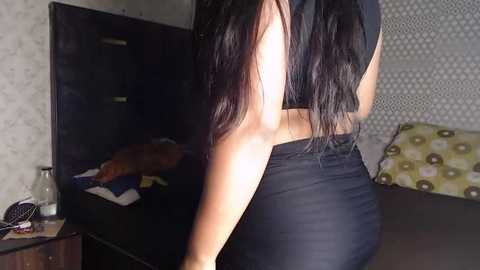 Media: Video of a woman with long black hair, wearing a black top and tight black shorts, standing in front of a TV and patterned wall, in a dimly lit room.