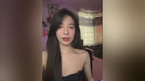 Media: A video of an East Asian woman with long black hair and fair skin, wearing a black strapless top, sitting on a bed in a dimly lit room with a window and curtains in the background.