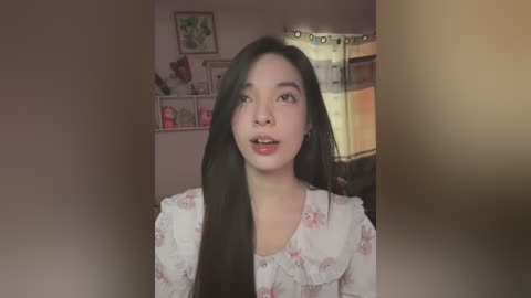 Media: Video of a young Asian woman with long black hair, wearing a white floral blouse, smiling in a softly lit, pink-walled bedroom with shelves and window curtains.