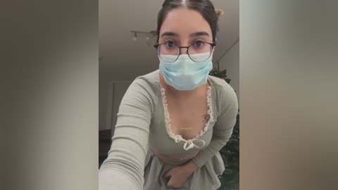 Media: Video of a young woman with light skin, wearing glasses, a light blue face mask, and a gray top with lace trim, bending over. Background features a blurred indoor setting with a Christmas tree.