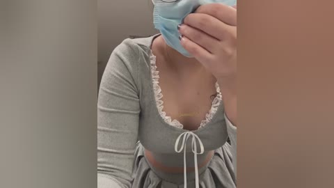 Video of a light-skinned woman in a gray, ruffled crop top with a blue mask, hand covering mouth, sitting on a beige surface, holding a phone.