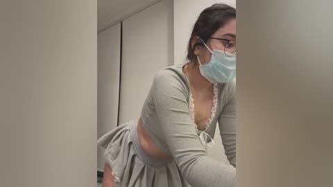 Media: A video of a young woman with medium skin tone, wearing a light gray crop top with a low V-neck, a blue surgical mask, and glasses, leaning against a white wall in a clinical setting.