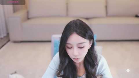 Video of a young Asian woman with long black hair, wearing a light blue cardigan, sitting on a light-colored floor, looking down with a serious expression. Background shows a beige sofa and a blue blanket.