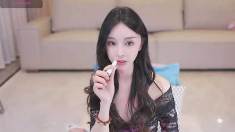 Media: Video of an East Asian woman with long black hair, fair skin, wearing a black lace top, applying makeup with a white stick in a modern living room with beige couches.