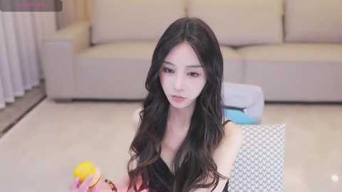 Media: Video of a young East Asian woman with long, wavy black hair, fair skin, and small breasts, seated on a light gray couch in a modern living room, holding a yellow ball.