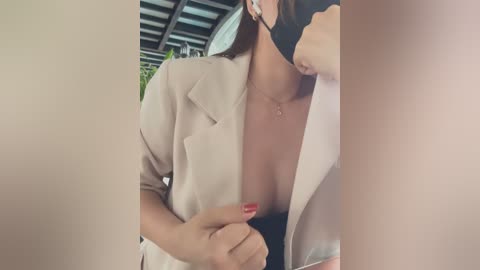 Media: Video of a woman with fair skin, wearing a beige blazer with a low neckline, revealing cleavage. She has long brown hair and is holding her phone, partially visible in the image.