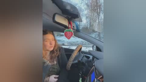 Media: Video of a woman with curly brown hair, wearing a green jacket, driving a car with a leopard-print steering wheel cover and a red Christmas ornament hanging from the rearview mirror, set against a snowy backdrop.