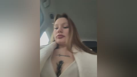 A video shows a white woman with long, straight brown hair, wearing a white blazer and a black top, seated in an airplane. She has a tattoo of cursive text on her chest.