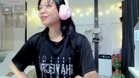 Media: Video of an East Asian woman with long black hair wearing a black T-shirt and pink headphones, standing in a cozy, dimly lit room with string lights, plants, and a white closet in the background.