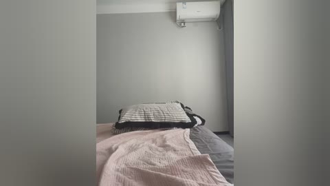 Media: Video of a minimalist, sparsely furnished room with a bed covered in a striped, black-and-white duvet, a pink blanket, and grey sheets. The walls are painted light grey, and an air conditioning unit is mounted on the wall above the bed.