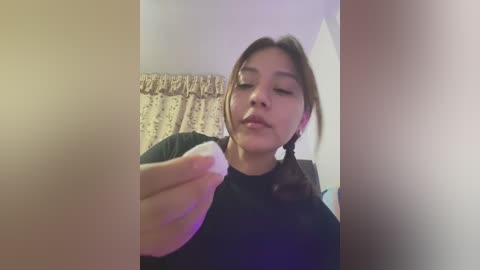 Media: Video of a young Asian woman with long brown hair, wearing a black top, holding a white object in front of her face, with floral curtains and a beige wall in the background.