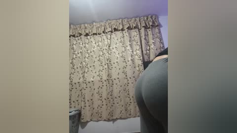 Media: Video of a woman's buttocks in tight gray leggings, partially visible, standing in front of a floral-patterned beige curtain in a dimly lit room.