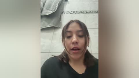 Media: Video of a young woman with medium skin tone and dark brown hair, wearing a black jacket, in a tiled bathroom with a towel hanging on the wall.