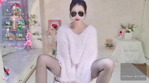 A video of a woman with fair skin, dark hair, and large sunglasses, sitting with legs spread, wearing a fluffy white sweater, black stockings, and sitting in a room with a pink background and gaming-themed decorations.