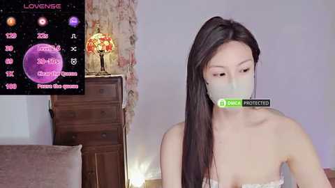 Media: A video of an Asian woman with long black hair, wearing a surgical mask, seated indoors with a wooden dresser and lamp in the background.