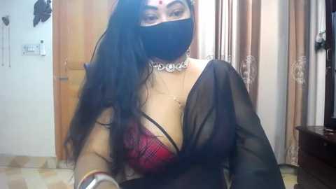 Media: Video of a woman with long black hair, wearing a black mask, red bra, and sheer black blouse. She has a large, ornate necklace. Background includes beige curtains and a wooden door.