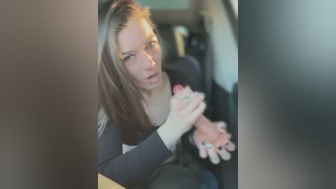 Media: Video of a young woman with long, straight brown hair and fair skin, wearing a black top, sitting in a car. She appears distressed, with her hands clasped near her face. The background features a blurred interior with dark upholstery.