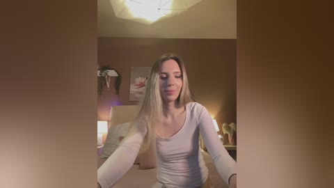 Media: Video of a fair-skinned, blonde woman with long hair, wearing a white long-sleeve top, sitting in a dimly lit bedroom with a beige wall and a lit lamp.